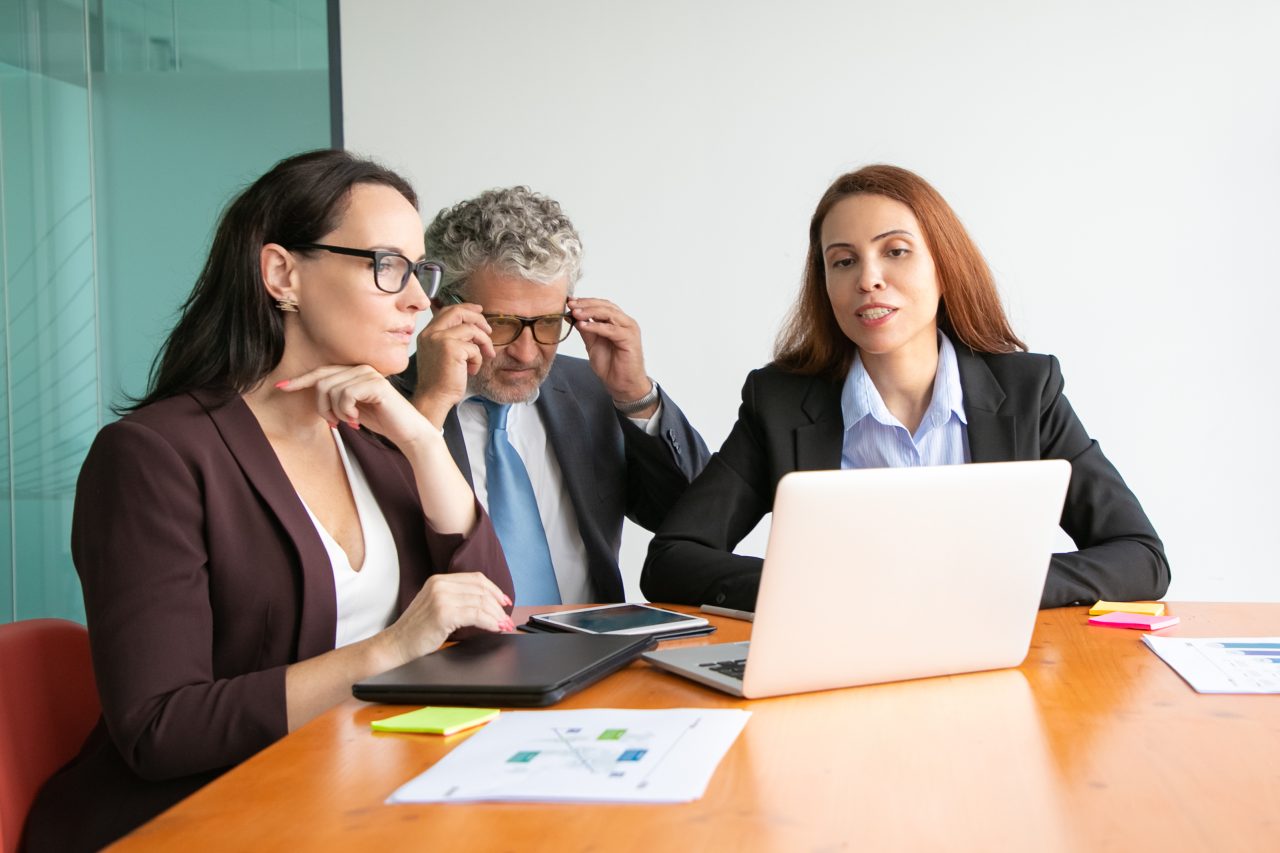 Business people watching project presentation on laptop, sitting at meeting table with paper reports and charts. Medium shot. Marketing and teamwork concept